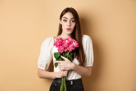 Valentines day. Image of surprised girlfriend thanking for flowers, receive pink roses from lover and looking grateful at lover, standing on beige background.