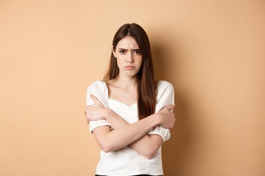 Offended woman hugging herself and frowning upset, feeling timid and defensive, standing on beige background.