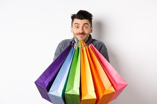 Excited young man carry colorful shopping bags and smiling happy, shopper buying on sale, standing on white background.