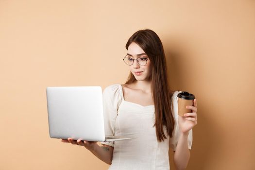 Portrait of woman working on laptop and drinking coffee. Girl student using computer for remote studies, standing on beige background.