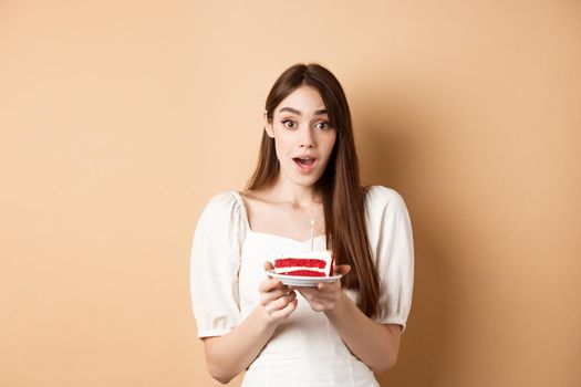Excited birthday girl looking amazed, holding cake with candle, making b-day wish, standing on beige background.