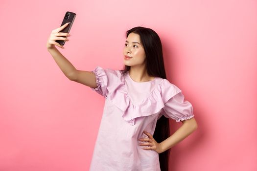 Stylish asian girl taking selfie and smiling, posing for social media photo, standing in dress against pink background.