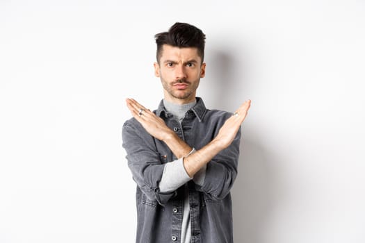 Angry man show cross gesture to stop or say no, frowning and looking serious, disagree and prohibit bad situation, standing on white background.