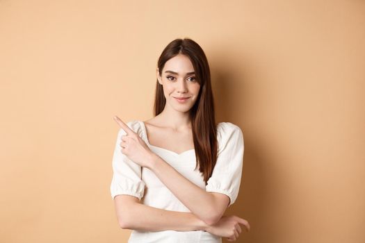 Smiling good-looking girl in white dress pointing aside, showing left logo and staring at camera confident, standing on beige background.