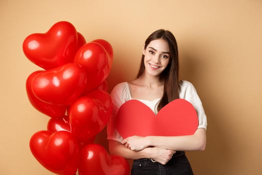 Valentines day and love concept. Lovely girl hugging big romantic heart cutout and smiling, standing near red balloons on beige background.