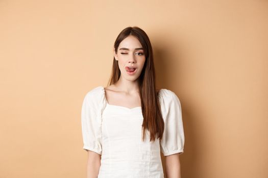 Cheeky young woman showing tongue and winking at camera, standing in white blouse on beige background.