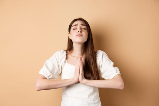 Hopeful woman close eyes and pray god, holding hands in begging gesture and making wish, say please, standing on beige background.