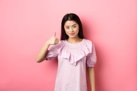 Smiling asian girl show thumb up in approval, praise good choice, like and recommend product, standing on pink background.