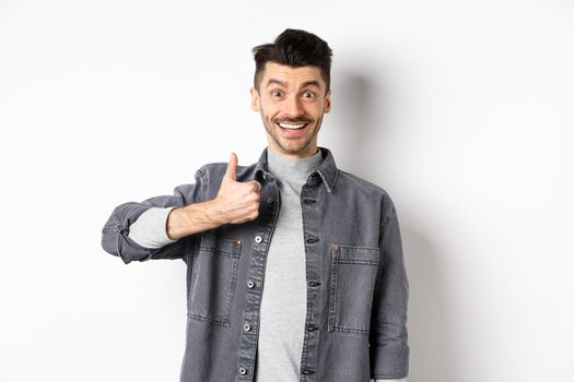 Excited handsome guy showing thumb up and smiling, like good thing, praise awesome choice, looking satisfied and happy, standing on white background.