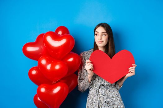 Valentines day. Romantic girl in dress standing near balloons and holding big red heart cutout, dream of something, standing on blue background.