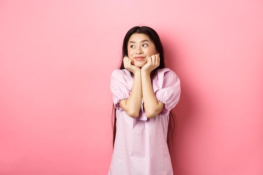 Dreamy romantic girl looking aside at logo and smiling, thinking of something beautiful, standing on pink background.