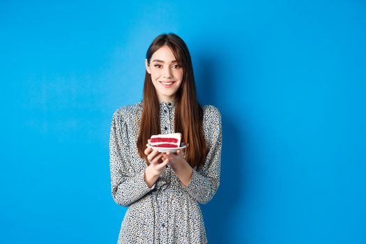 Beautiful girl holding birthday cake and celebrating, wishing happy bday and smiling, standing against blue background.