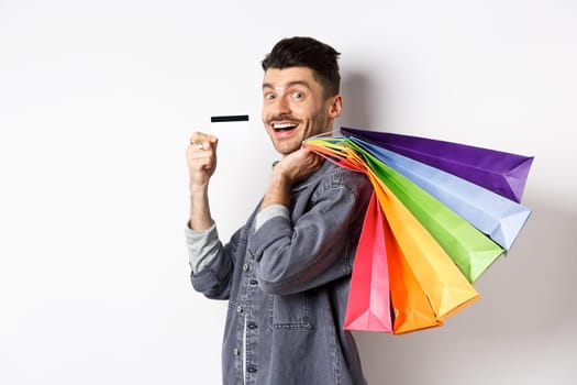Cheerful guy going on shopping with credit card, holding bags on shoulder and smiling excited at camera, white background.