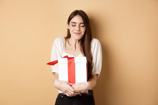Happy valentines day. Dreamy woman hugging her surprise gift, close eyes and smile, standing on beige background.