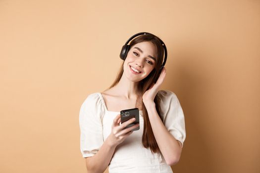 Cute smiling girl listening music in wireless headphones, using smartphone, standing on beige background.