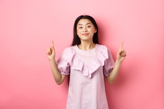 Beautiful young asian woman in romantic dress pointing fingers up, smiling and gladly showing advertisement, standing on pink background.