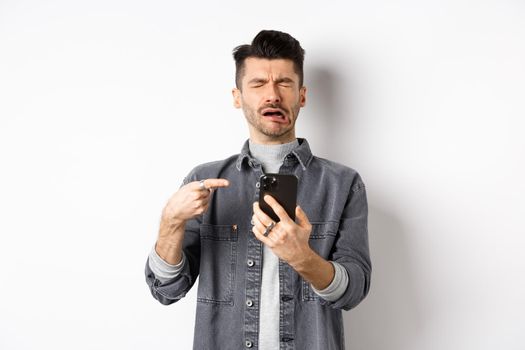 Sad crying guy pointing at smartphone and sobbing, complaining or feeling miserable, standing on white background.