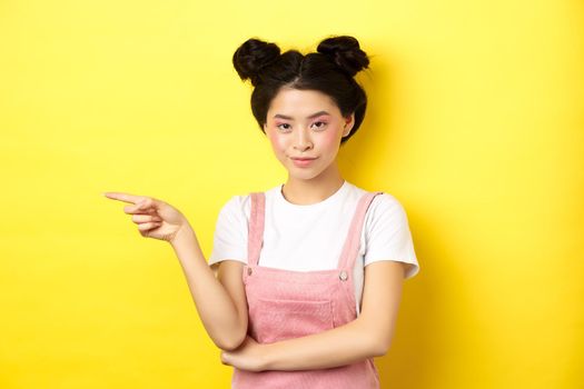 Beautiful teenage asian girl with bright makeup, pointing finger left at banner and smiling, yellow background.