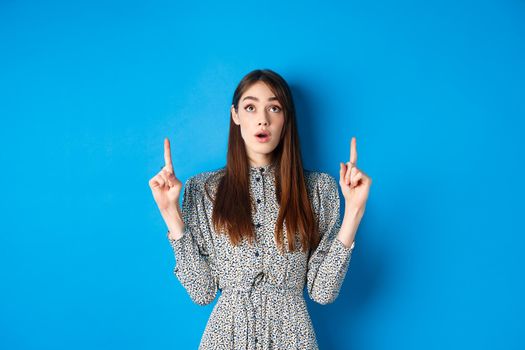 Surprised romantic girl in dress pointing, looking up with opened mouth and fascinated gaze, standing on blue background.