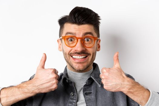 Supportive young man in glasses smiling, showing thumbs up to praise or approve good thing, standing against white background.