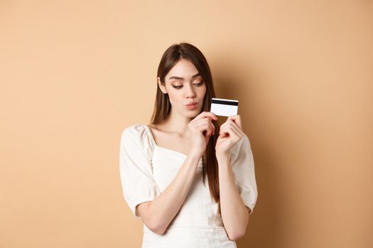 Cute young woman looking thoughtful at plastic credit card, thinking of shopping, standing on beige background.