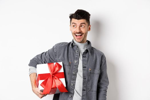 Cheerful handsome guy looking at empty space with amazed smile, checking out special valentines day offer, holding big romantic gift box, standing on white background.