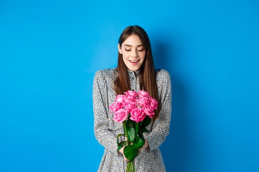 Valentines day concept. Happy attractive woman receive surprise flowers, looking thankful at bouquet of pink roses, standing on blue background.