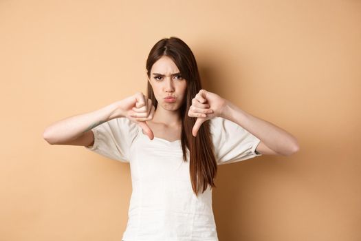 Absolutely no. Disappointed young woman frowning and showing thumbs down, express dislike and negative emotions, bad feedback, standing on beige background.