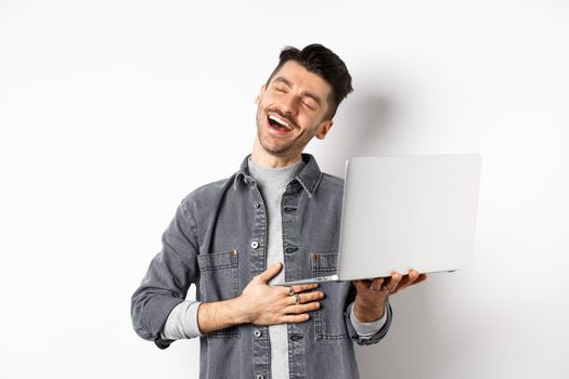 Image of happy guy laughing at funny video on laptop, touching belly and smiling satisfied wth good laugh, standing against white background.