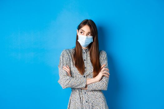 Covid-19, pandemic lifestyle concept. Pensive girl in medical mask look at upper left corner logo, thinking ways of protecting health from coronavirus, blue background.