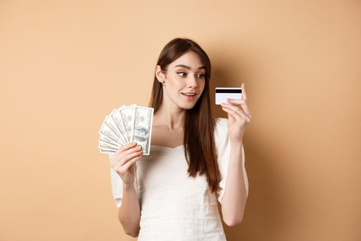 Dreamy girl looking at plastic credit card and thinking of shopping, holding dollar bills, standing on beige background.