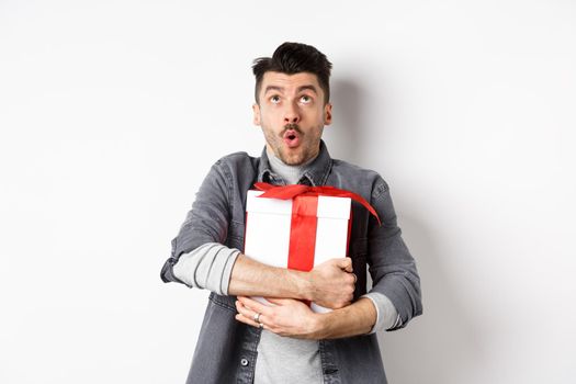 Surprised funny guy hugging big holiday gift and looking up, say wow amazed, checking out Valentines day special offer, standing on white background.