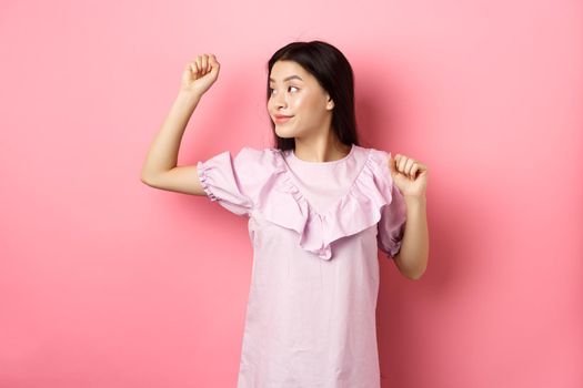 Carefree asian girl dancing, raising hand up and looking left at logo, standing in romantic dress against pink background.