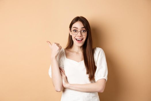 Excited girl in glasses pointing aside at logo, smiling amazed, recommending company, standing on beige background.