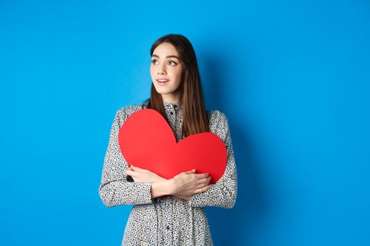 Valentines day. Hopeful girl dreaming off soulmate, looking aside at empty space with excitement, hugging big red heart cutout, standing on blue background.