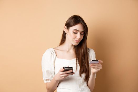 Smiling young woman paying online, looking at credit card and making purchase on mobile phone, shopping in internet, standing on beige background.