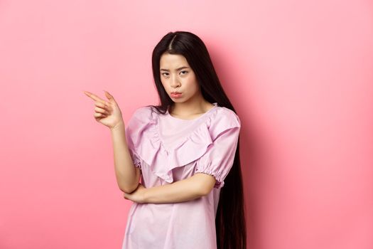 Sad and grumpy asian girl sulking, pointing left and look at camera jealous, want something, complaining and showing desired thing, standing on pink background.