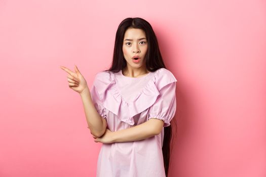 Shocked asian teen girl in dress pointing finger left, gasping wondered and look curious at camera, asking question about promo, standing on pink background.