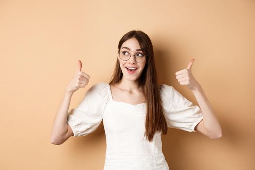 Happy girl in glasses show thumbs up, looking aside at good logo, standing satisfied on beige background.