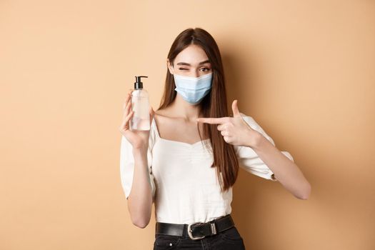 Covid-19 and preventive measures concept. Smiling girl winking in medical mask, pointing at hand sanitizer bottle, showing good product, beige background.