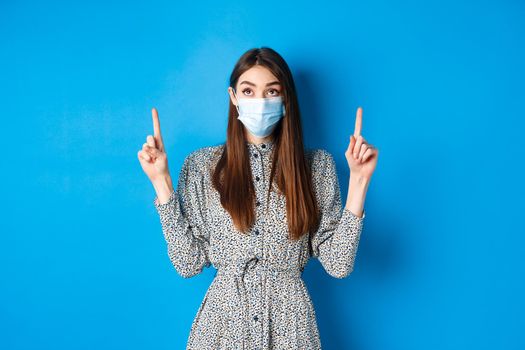 People, covid and quarantine concept. Intrigued young woman looking and pointing up at advertisement, standing in dress and face mask from coronavirus, blue background.