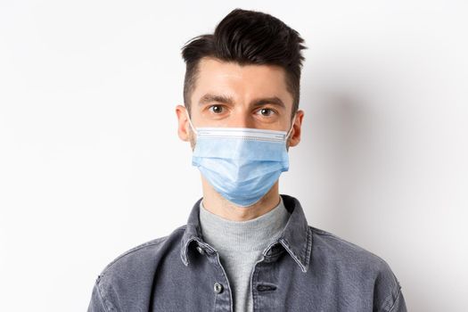 Pandemic lifestyle, healthcare and medicine concept. Close up of candid young man wearing medical mask from covid-19, taking care of health, standing on white background.