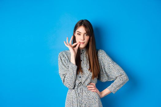 Serious woman promise to keep secret, seal lips, making zip gesture and looking at camera, standing in dress on blue background.