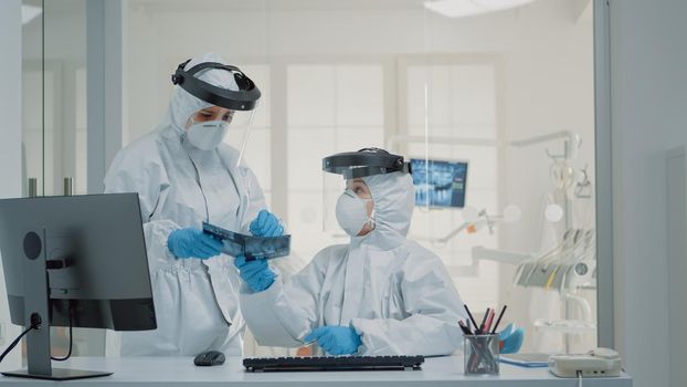 Medical stomatology staff working on teeth healthcare for patient at dental clinic, wearing protection suits. Nurse using modern computer at desk while dentist holding x ray for implant