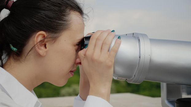 Close up of young woman using binoculars for sightseeing on top of skyline building. Caucasian person with telescope watching outdoor urban scene from observation point on tower