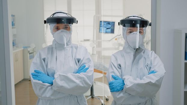 Stomatological staff standing at dental clinic while wearing protection suit with coverall, gloves, face shield and mask during covid pandemic. Team of dentists looking at camera