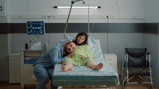 Caucasian couple expecting child in hospital ward while african american nurse preparing IV drip bag for pregnant woman at maternity clinic. Husband and wife waiting for baby delivery