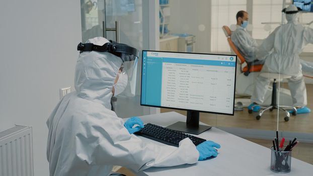 Stomatology nurse sitting at desk using modern computer, wearing ppe suit. Dental assistant looking at monitor typing on keyboard and orthodontist with oral care equipment examining patient