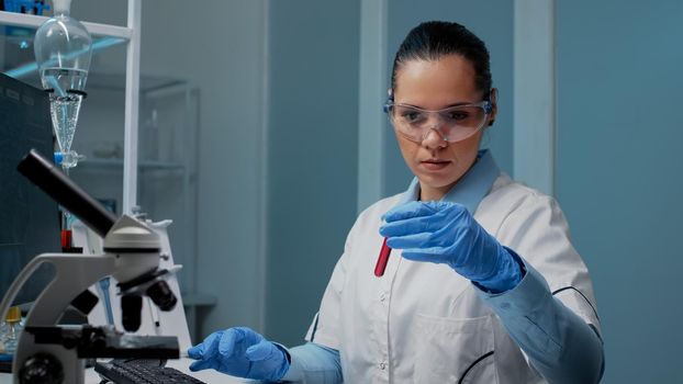 Microbiology doctor looking at vacutainer from tray with blood sample on desk and using computer in chemical laboratory. Research specialist doing healthcare experiment with dna glass flask