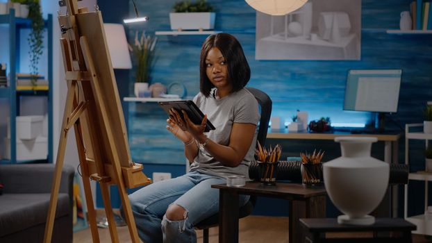 Woman of african american ethnicity holding digital tablet for creative drawing while sitting in workshop room at home. Black artist using technology for masterpiece inspiration